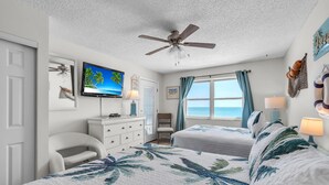 A bedroom with two beds, a ceiling fan, a TV, a dresser, and a view of the ocean. - Sea Breeze 503 (Madeira Beach)