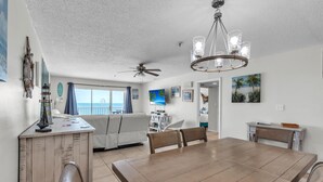 A spacious dining area with a wooden table and chairs, a ceiling fan, and a view of the ocean through large windows. - Sea Breeze 503 (Madeira Beach)
