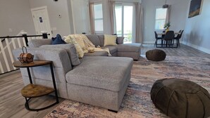 A modern living room with a grey sectional sofa, a small wooden side table, a brown pouf, and a dining area with a table and chairs. - 3BR Gulf Getaway | Elevator, Pool, Walk to IRB (Indian Rocks Beach)