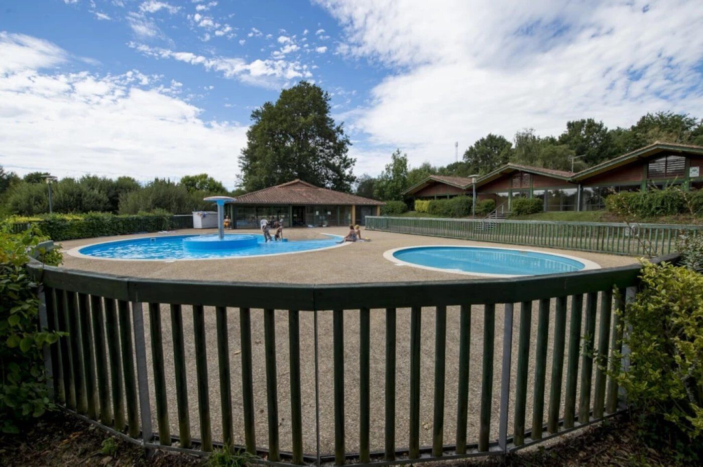 A fenced swimming pool area with two pools, surrounded by a wooden deck and a building with a tiled roof in the background.