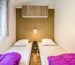 A small, compact room with two single beds, a wooden headboard, a small table, and a wall-mounted light fixture.