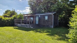 A small wooden cabin with a porch and a single door, surrounded by a grassy yard and trees.
