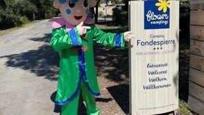 Une mascotte vêtue d’un costume vert et d’un chapeau rose se tient à côté d’un panneau indiquant « Flower Campings ».