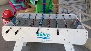 A foosball table with a "Tahiti Resort" logo, a mascot in a colorful outfit, and a covered outdoor area with chairs and a table. - ZEcamping 3 étoiles familial toboggans locatif TV clim 26m2 4 pers (Sussargues)