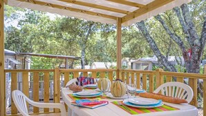 A covered outdoor dining area with a wooden deck, white chairs, and a table set for a meal. - ZEcamping 3 étoiles familial toboggans locatif TV clim 26m2 4 pers (Sussargues)
