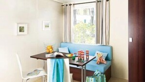 A compact room with a dining table, chairs, a sofa, and a window with curtains. - ZEcamping 3 étoiles familial toboggans locatif TV clim 26m2 4 pers (Sussargues)