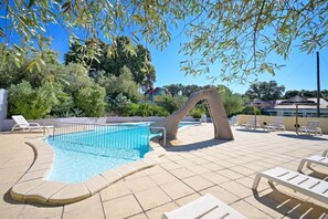 A swimming pool with a slide, surrounded by lounge chairs and umbrellas, with trees and buildings in the background.