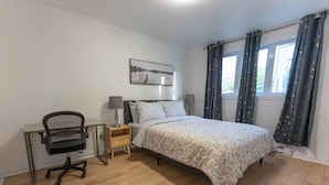 Ein Schlafzimmer mit einem Bett, einem Schreibtisch mit Stuhl, einer Lampe und einem Fenster mit Vorhängen.