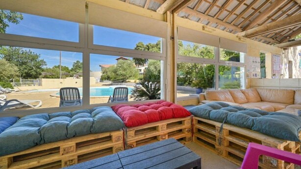 A covered outdoor seating area with wooden pallets, cushioned benches, and a view of a swimming pool and greenery. - ZEcamping 3 étoiles toboggans locatif TV LV clim 28m2 (Sussargues)