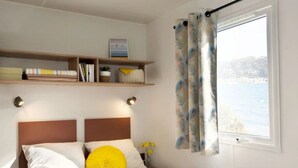 A neatly made bed with a white duvet and a yellow pillow, a bedside table with a lamp, and a window with curtains. - ZEcamping 3 étoiles toboggans locatif TV LV clim 28m2 (Sussargues)