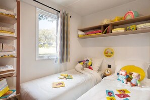 A child's bedroom with two beds, a window with curtains, and shelves with toys and books. - ZEcamping 3 étoiles toboggans locatif TV LV clim 28m2 (Sussargues)