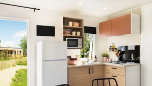A modern kitchen with a refrigerator, microwave, and stove. There is a dining table with chairs and a view of a garden outside. - ZEcamping 3 étoiles toboggans locatif TV LV clim 28m2 (Sussargues)