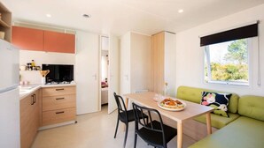 A compact kitchen with a dining area, a green sofa, and a window with a view of greenery. - ZEcamping 3 étoiles toboggans locatif TV LV clim 28m2 (Sussargues)