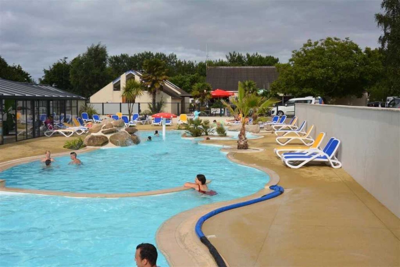 En swimmingpool med flere baner, omgivet af liggestole og parasoller, og en bygning i baggrunden.
