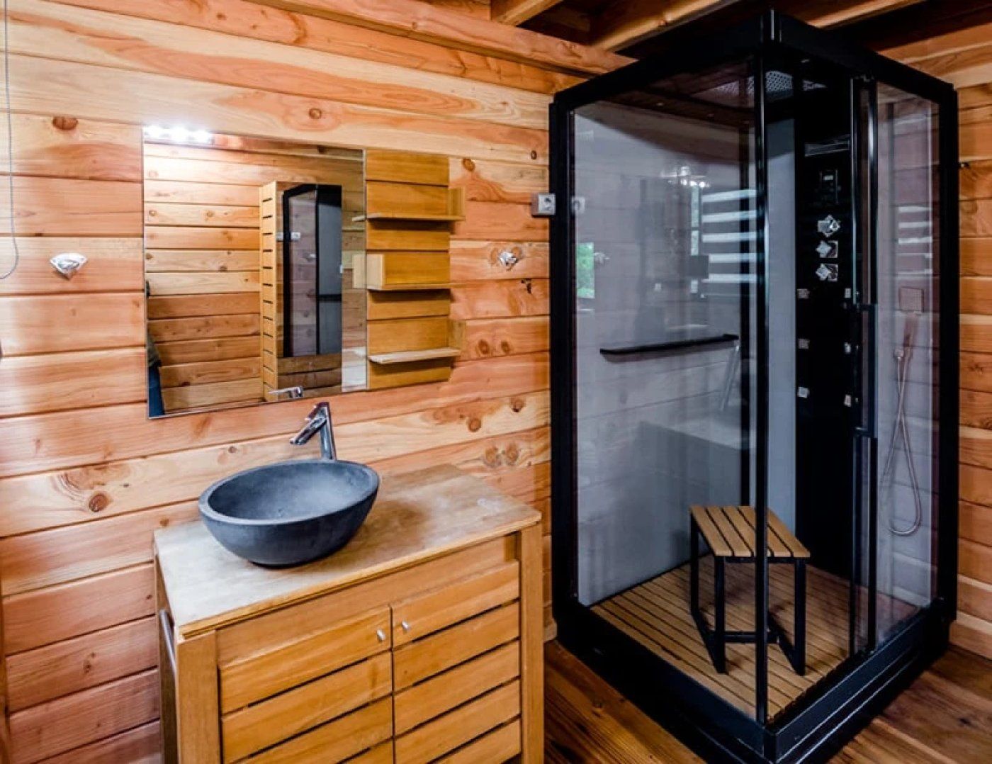 A wooden cabin interior with a wooden vanity, a bowl sink, a mirror, and a glass-enclosed shower area.