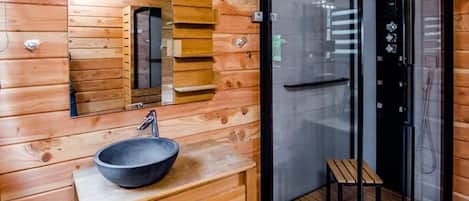 Interior de una cabaña de madera con tocador de madera, lavabo tipo cuenco, espejo y zona de ducha con mampara de vidrio.