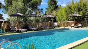 Ein Swimmingpool mit Liegestühlen und Strohdächern, umgeben von einem Holzzaun und Bäumen.