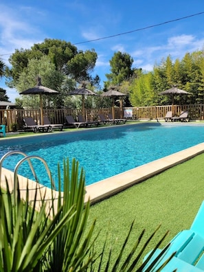 A swimming pool with lounge chairs and thatched umbrellas, surrounded by a wooden fence and greenery.