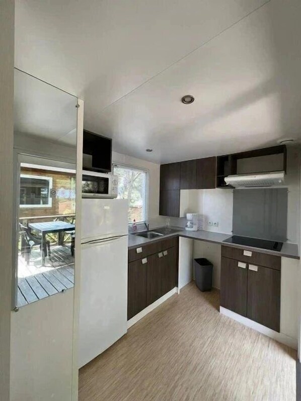 A modern kitchen with dark wood cabinets, a stainless steel sink, and a built-in oven. There is a trash bin and a window overlooking a deck area.