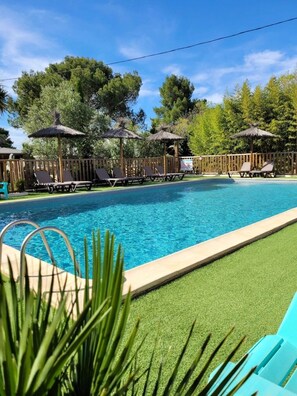 Une piscine entourée d’une clôture en bois et de verdure, agrémentée de chaises longues et de parasols en chaume.
