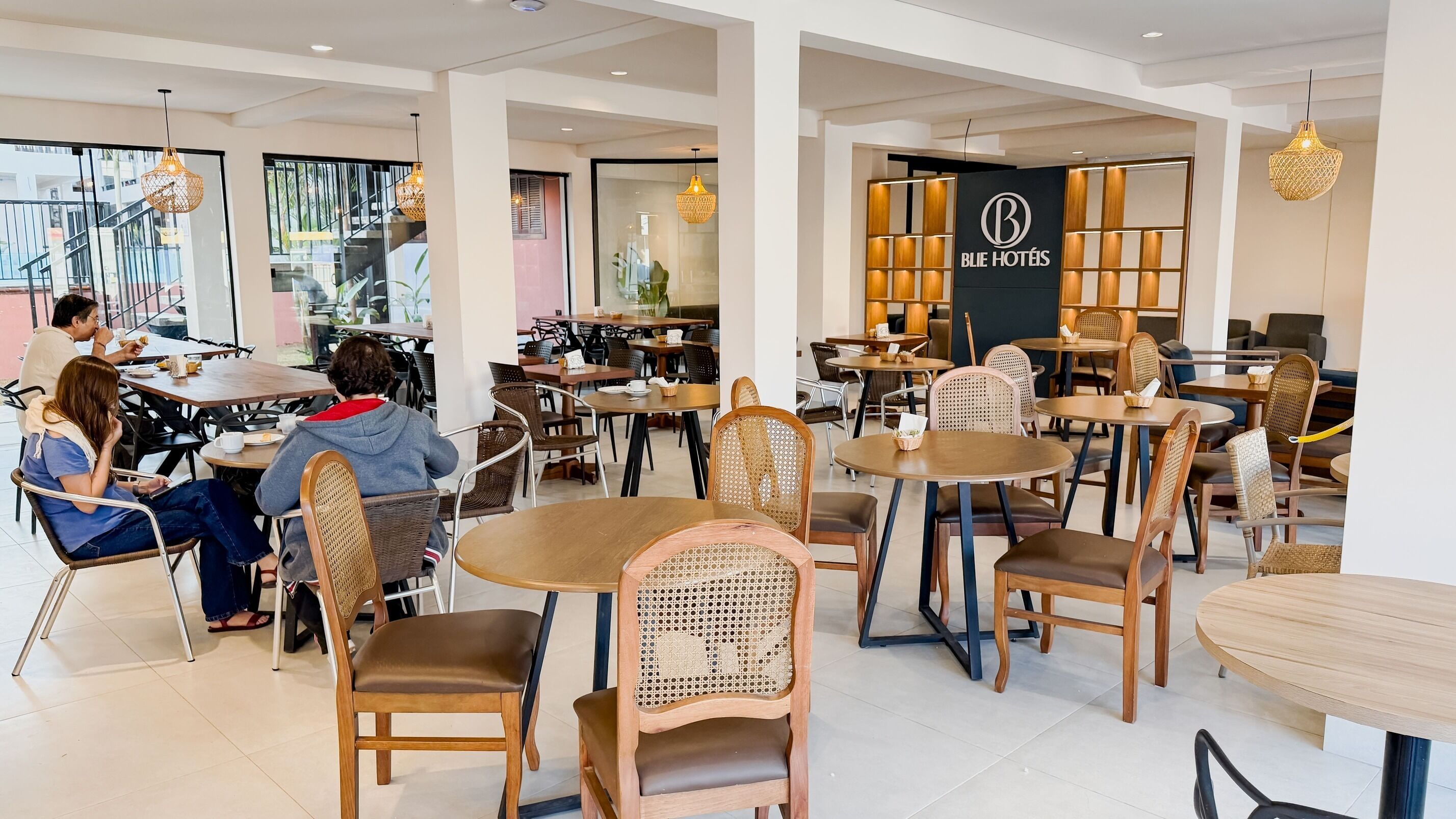 Un interno moderno di un hotel con tavoli rotondi, sedie in vimini e un cartello visibile per 'BLIE HOTÉIS'.