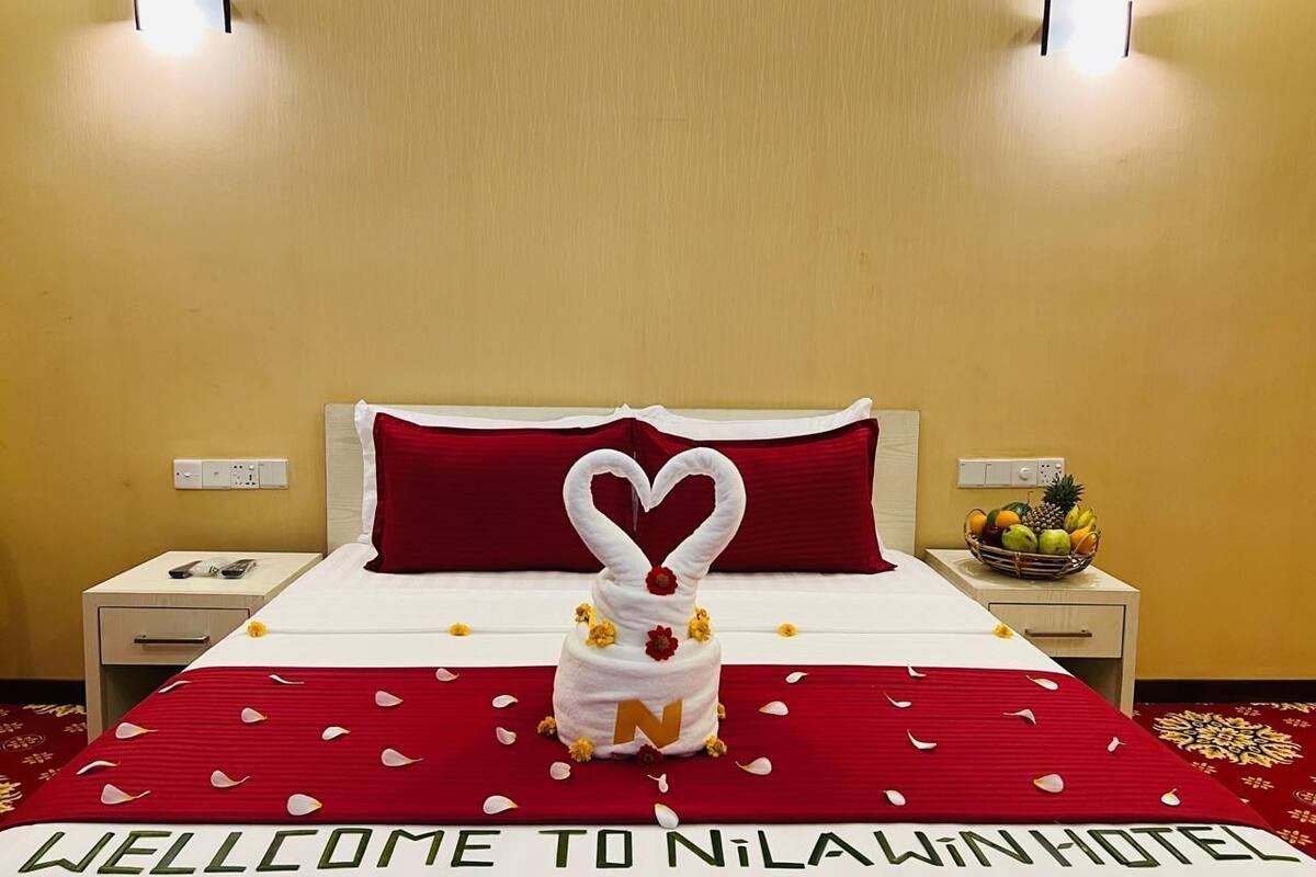 Une chambre d’hôtel avec un lit, une couvre-lit rouge et blanche, une décoration en forme de cœur faite avec une serviette, un panier de fruits et un panneau de bienvenue.