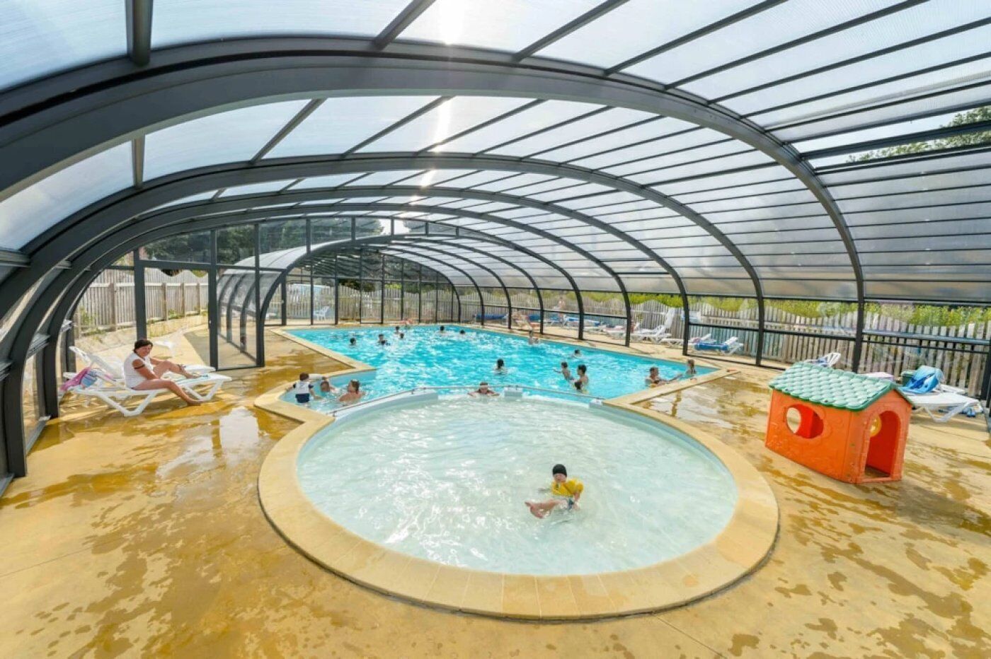 En indendørs swimmingpool med en buet tagkonstruktion, omgivet af liggestole og et lille legeområde.