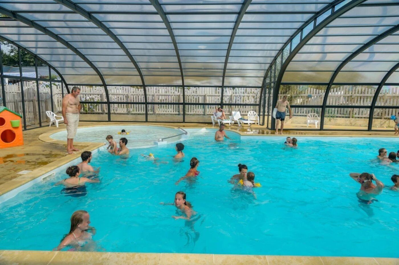 En pool med tak över, där folk badar och leker, omgiven av ett staket och ett trädäck.