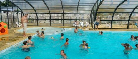 Una piscina techada con gente nadando y jugando, rodeada por una cerca y una terraza de madera.