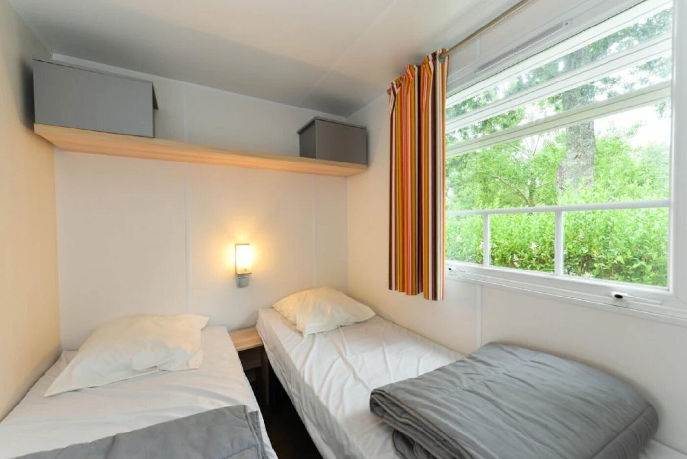 Una habitación pequeña y limpia con dos camas, un estante encima de ellas, una lámpara de pared y una ventana con cortinas.
