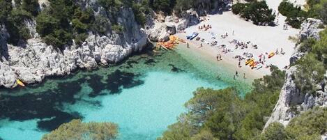 A coastal beach area with rocky cliffs, clear turquoise waters, and a sandy beach with people and boats.