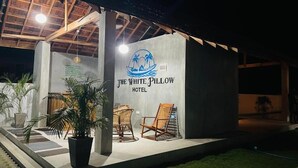 A building with a sign reading 'The White Pillow Hotel' and a logo with palm trees and a boat. There are two wicker chairs outside.