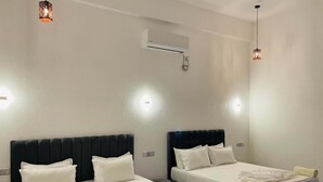 A hotel room with two beds, white linens, a black headboard, a wall-mounted air conditioner, and two pendant lights.