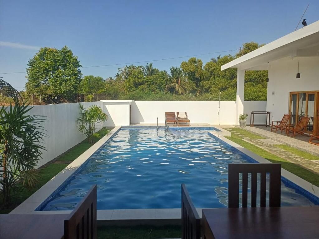 A rectangular swimming pool with a wooden deck in the foreground, a bench, and a white building with a balcony in the background.