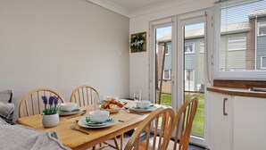 Ein Essbereich mit einem Holztisch, gedeckt für vier Personen, umgeben von Holzstühlen. Auf dem Tisch steht ein kleiner Blumentopf, und zur Rechten befindet sich ein Fenster mit Jalousien.