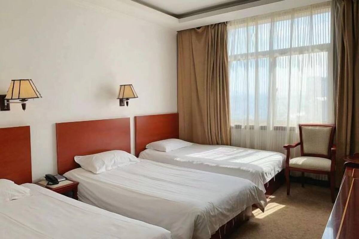 Une chambre d’hôtel avec trois lits simples, une tête de lit en bois, une chaise et un bureau avec une lampe.