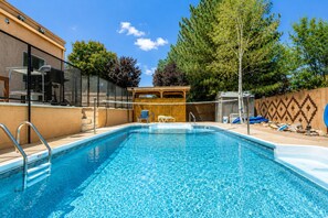 Ein klares, blaues Schwimmbecken mit einer Holzterrasse und Liegestühlen, umgeben von einem Zaun und Bäumen.