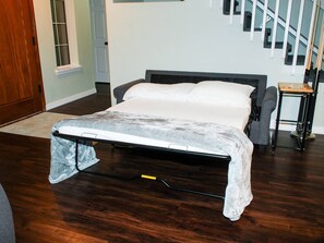 Una cama bien hecha en una habitación con piso de madera, una escalera al fondo y una pequeña mesa con una silla.