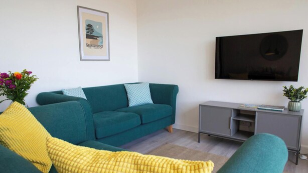 A living room with a teal sofa, yellow throw pillow, a framed picture on the wall, a grey TV stand, and a small plant. - Gweld y Mor, Saundersfoot (Saundersfoot)