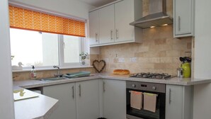 A kitchen with white cabinets, a gas stove, an oven, a window with an orange patterned blind, and a heart-shaped decoration on the wall. - Gweld y Mor, Saundersfoot (Saundersfoot)