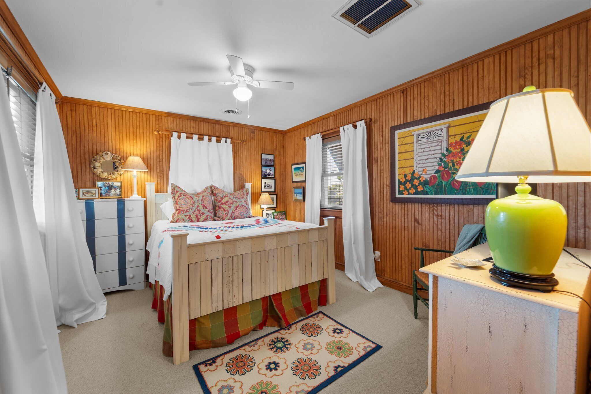 A bedroom with a bed, a dresser, a desk, a lamp, a ceiling fan, and a window with curtains.