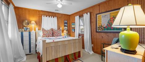 A bedroom with a bed, a dresser, a desk, a lamp, a ceiling fan, and a window with curtains.