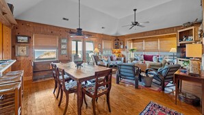 A spacious living area with a wooden dining table, comfortable seating, and a large window offering a view. - 'Loophole' | Oceanfront | Historic Cottage (Nags Head)