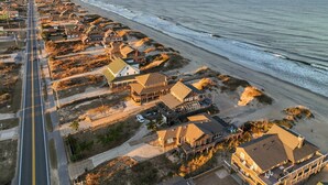 An aerial view of a coastal residential area with houses along the beach, a road, and a pier in the distance. - 'Loophole' | Oceanfront | Historic Cottage (Nags Head)