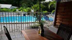 Una mesa de madera con dos sillas, una botella y una piña en un balcón con vista a una piscina.