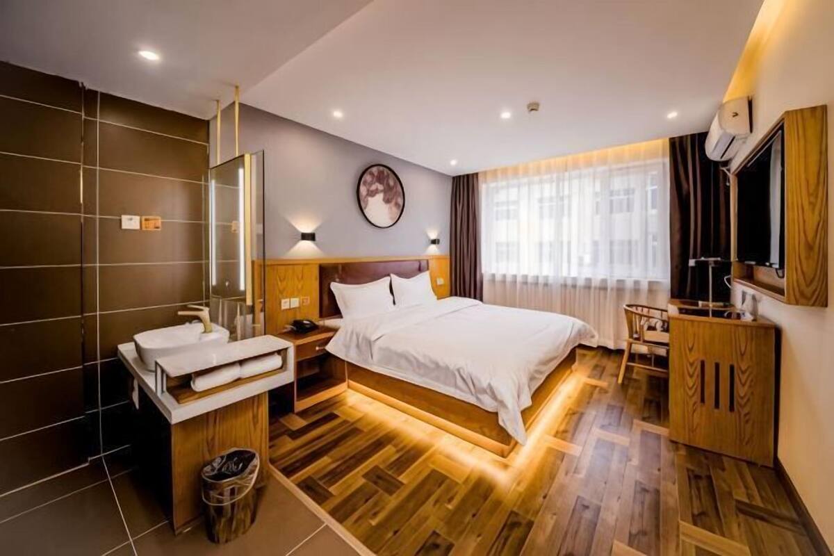A modern hotel room with a wooden floor, a bed with white linens, a wooden nightstand, a mirror, a wall-mounted TV, and a window with curtains.