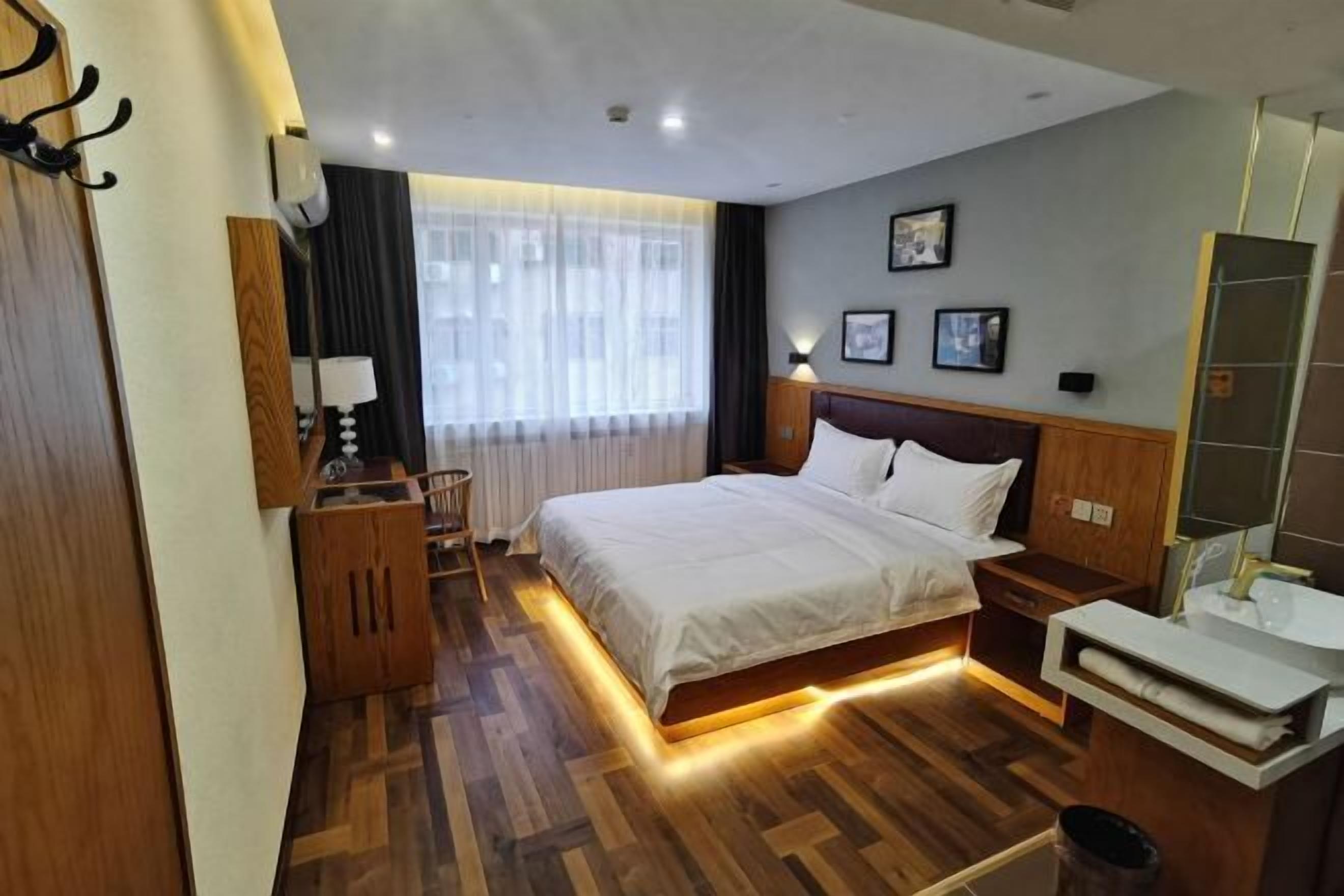 A hotel room with a wooden floor, a bed with white bedding, a bedside table with a lamp, a small wooden cabinet, a mirror, and a window with sheer curtains.