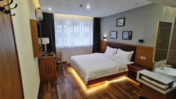 A hotel room with a wooden floor, a bed with white bedding, a bedside table with a lamp, a small wooden cabinet, a mirror, and a window with sheer curtains.