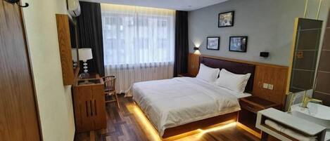 A hotel room with a wooden floor, a bed with white bedding, a bedside table with a lamp, a small wooden cabinet, a mirror, and a window with sheer curtains.