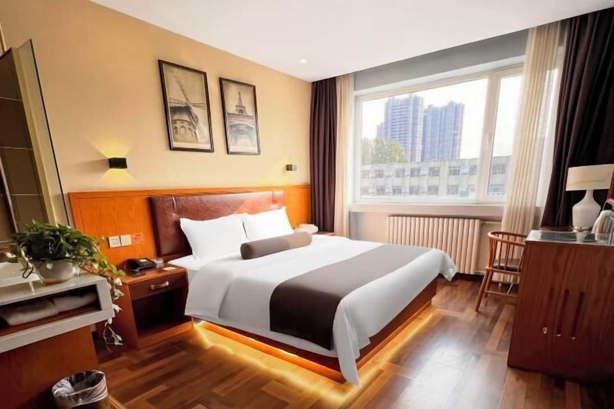 A hotel room with a large bed, a nightstand, a desk, a chair, and a window with a view of buildings.